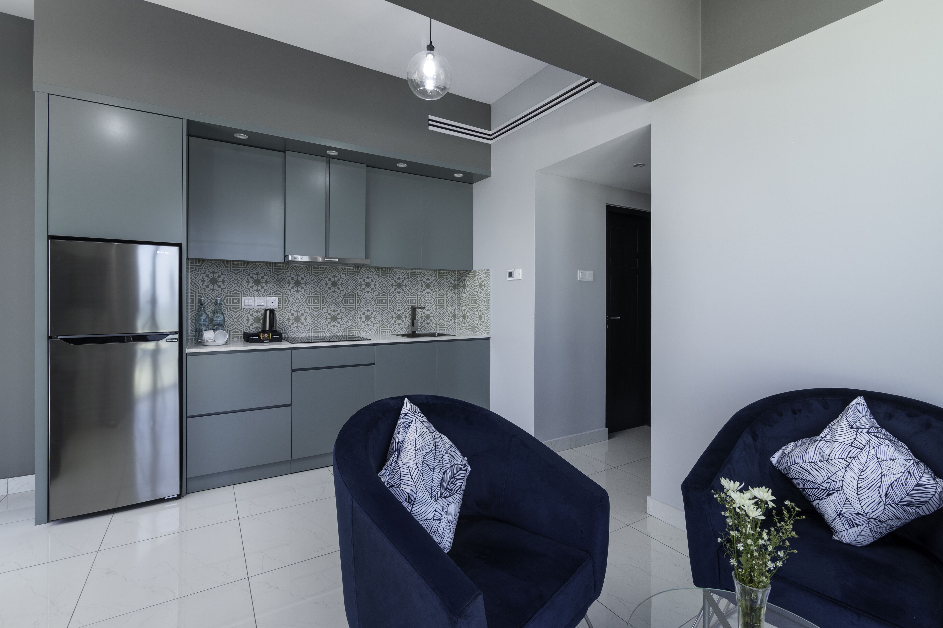A modern kitchen with grey cabinets, a stainless steel refrigerator, and a patterned backsplash. There are two dark blue armchairs with patterned cushions, a small table with a vase of flowers, and a hanging light fixture.