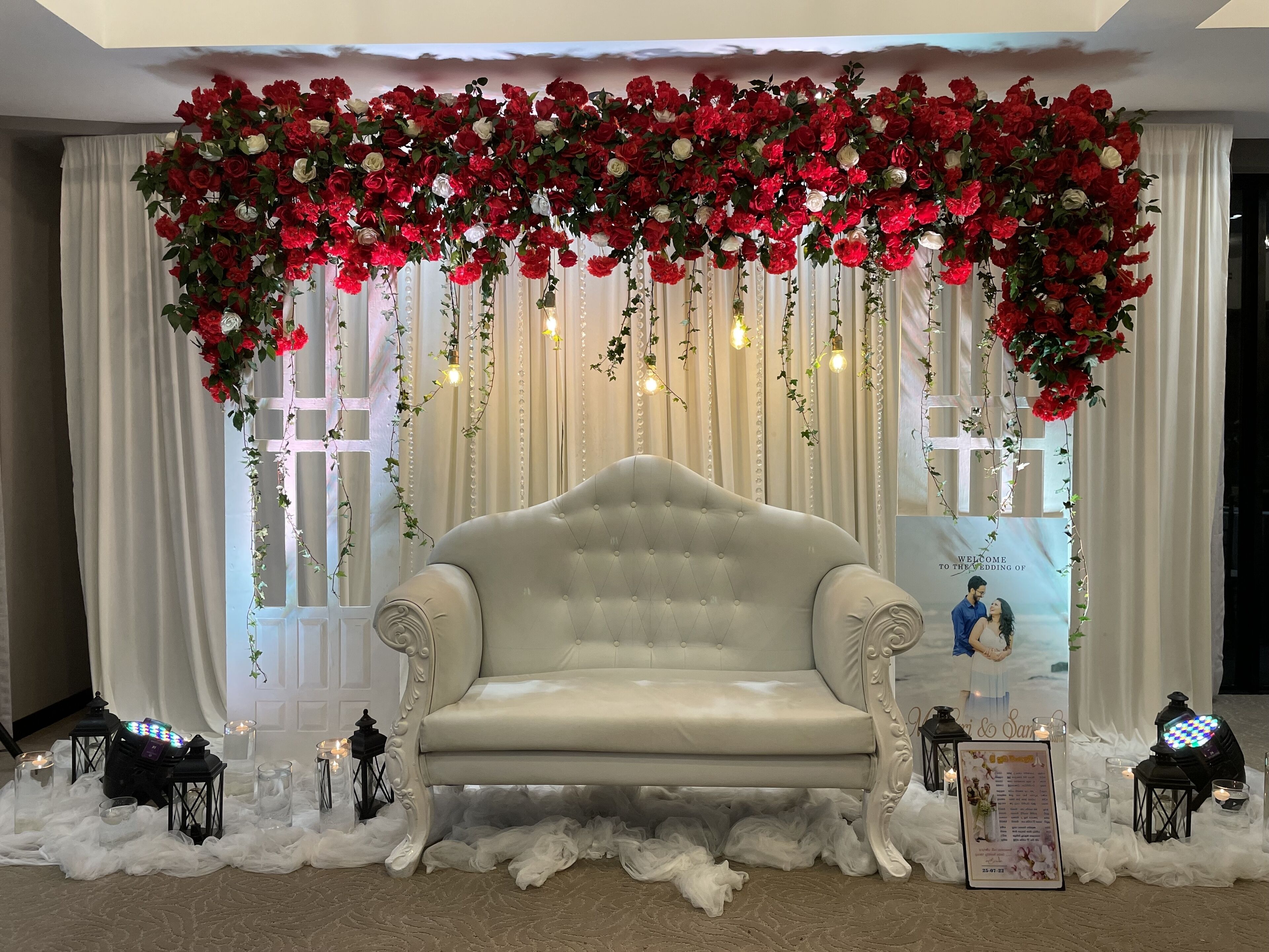 A stage with a white sofa, red and white flowers, hanging lights, and a backdrop with a wedding invitation.