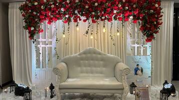 A stage with a white sofa, red and white flowers, hanging lights, and a backdrop with a wedding invitation.