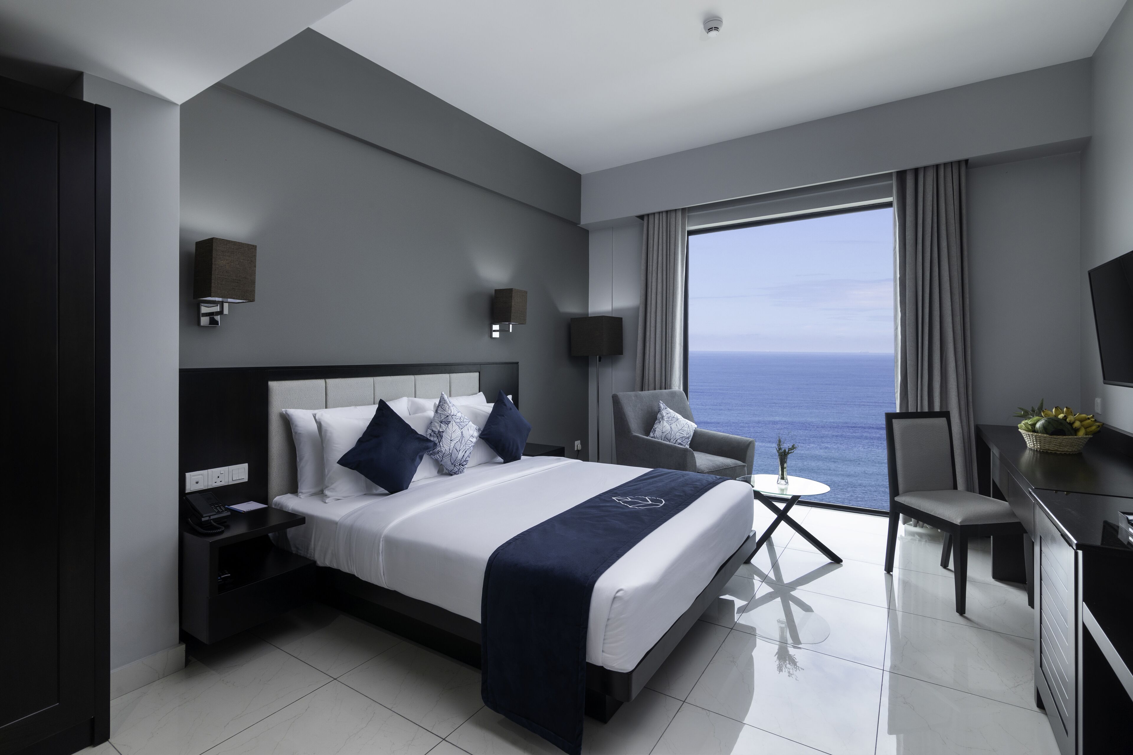 A modern hotel room with a large bed, a desk, a chair, and a view of the ocean.
