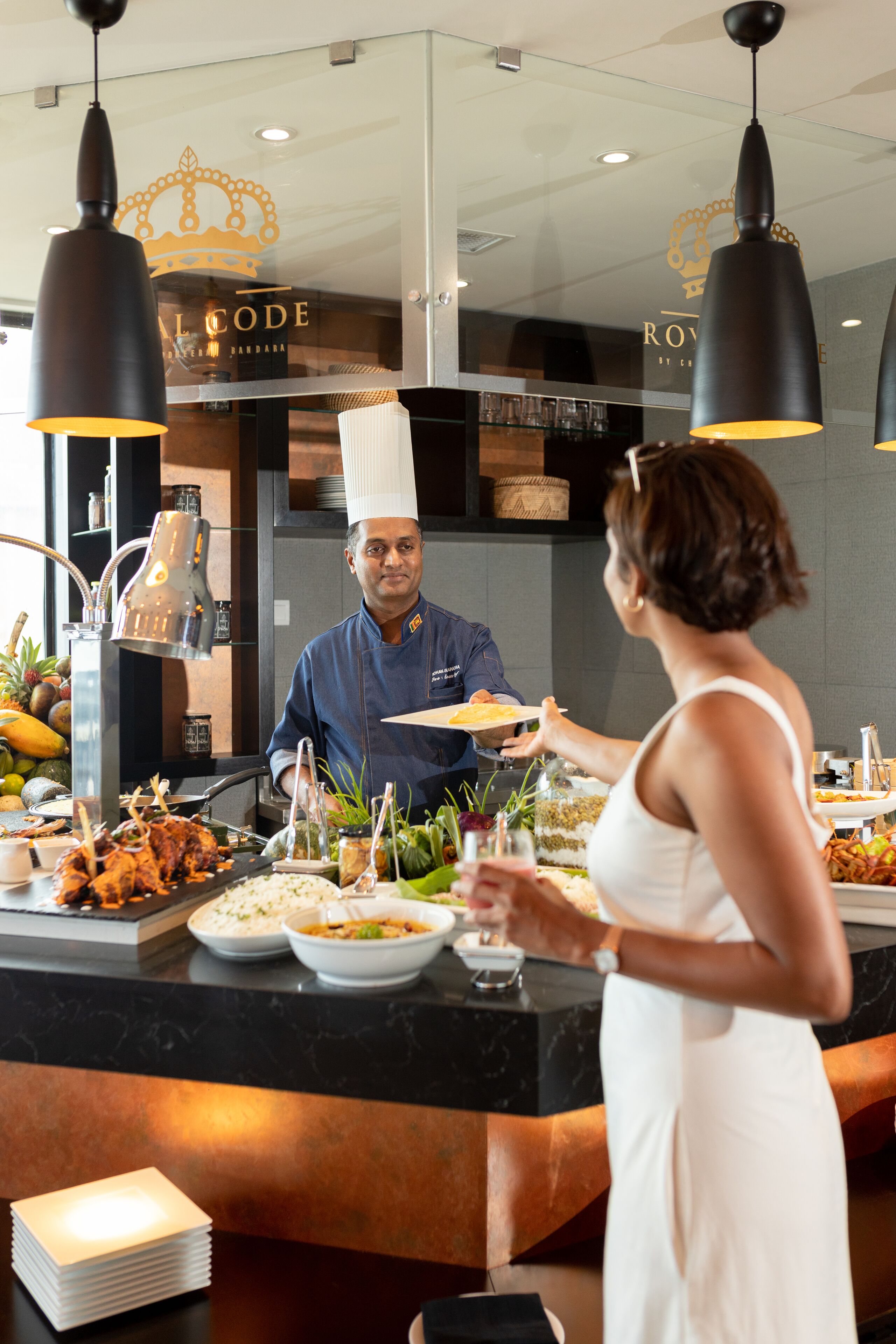 A buffet setup with a chef serving food, a woman holding a plate, and various dishes and drinks on the counter.