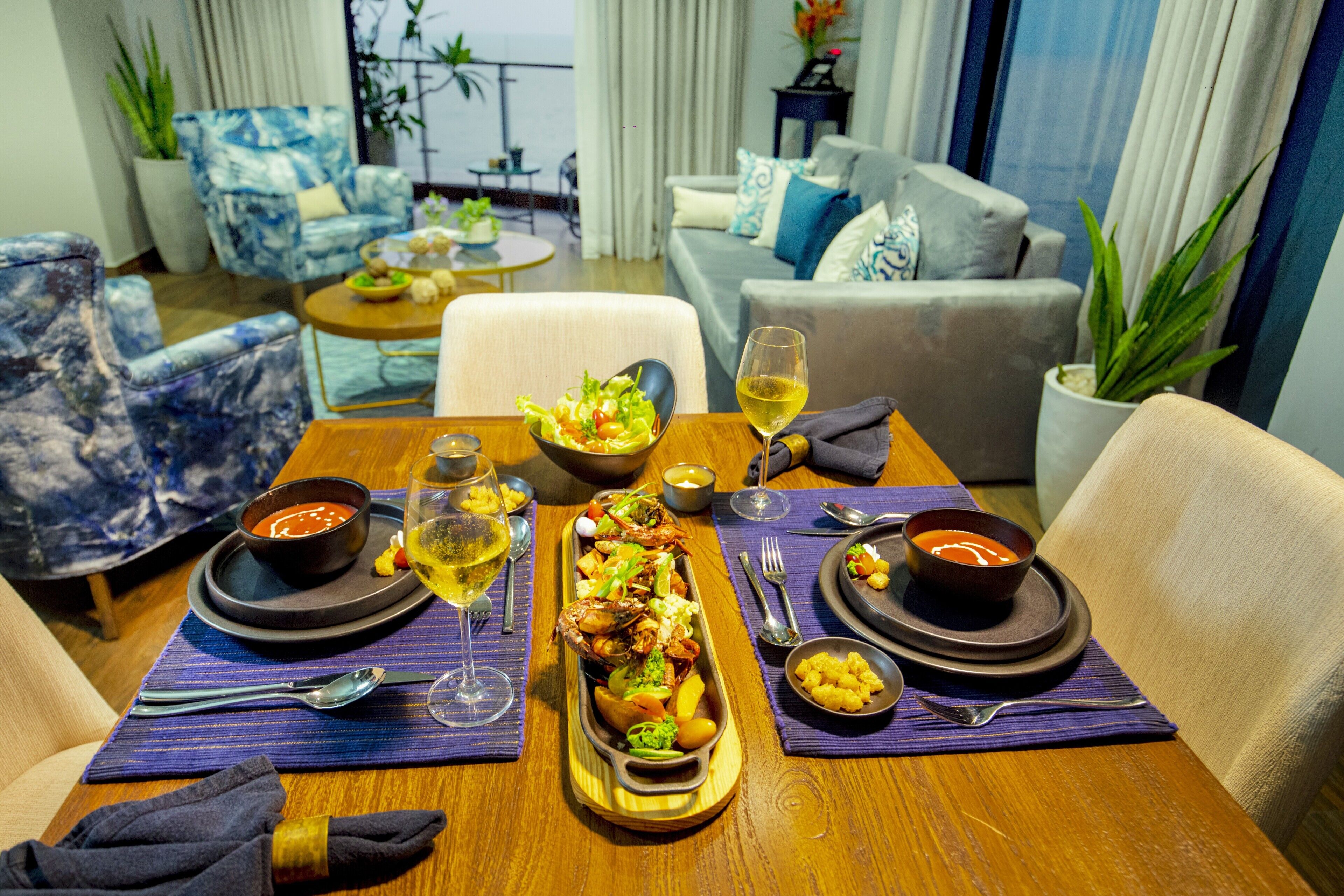Una mesa de comedor servida con un plato de mariscos, tazones de sopa y una copa de vino, con vista al mar a través de amplias ventanas.