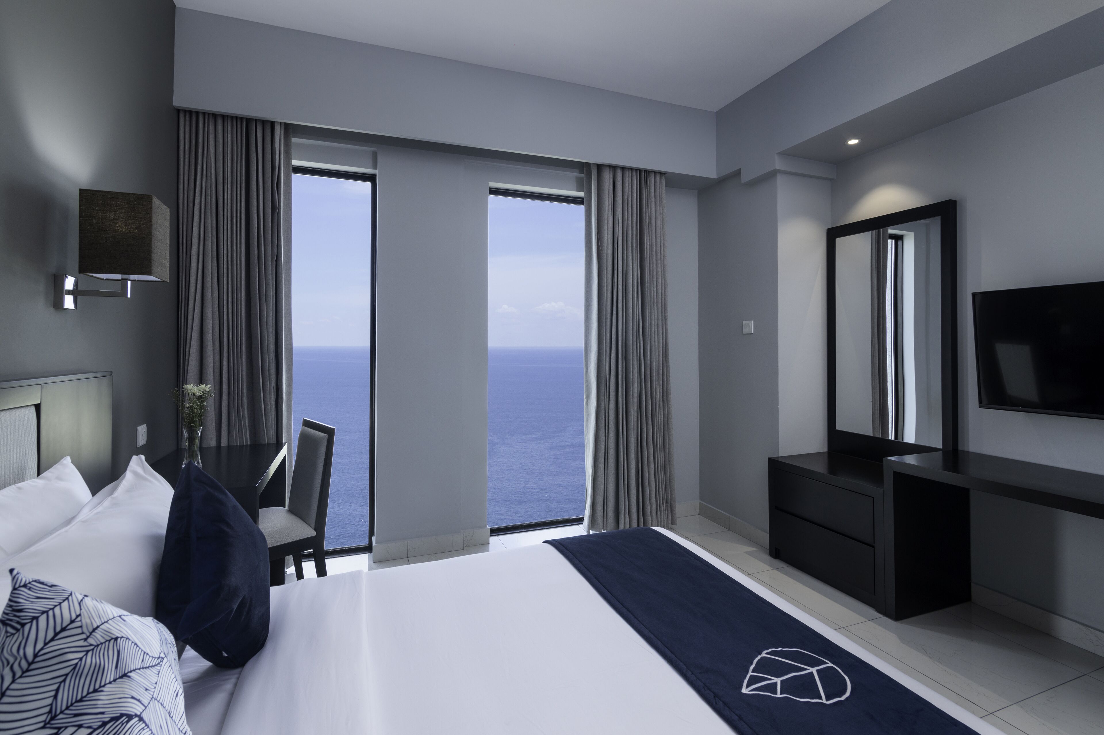 A modern hotel room with a large bed, a desk with a chair, a TV, and a view of the ocean.