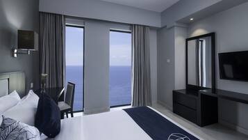 A modern hotel room with a large bed, a desk with a chair, a TV, and a view of the ocean.