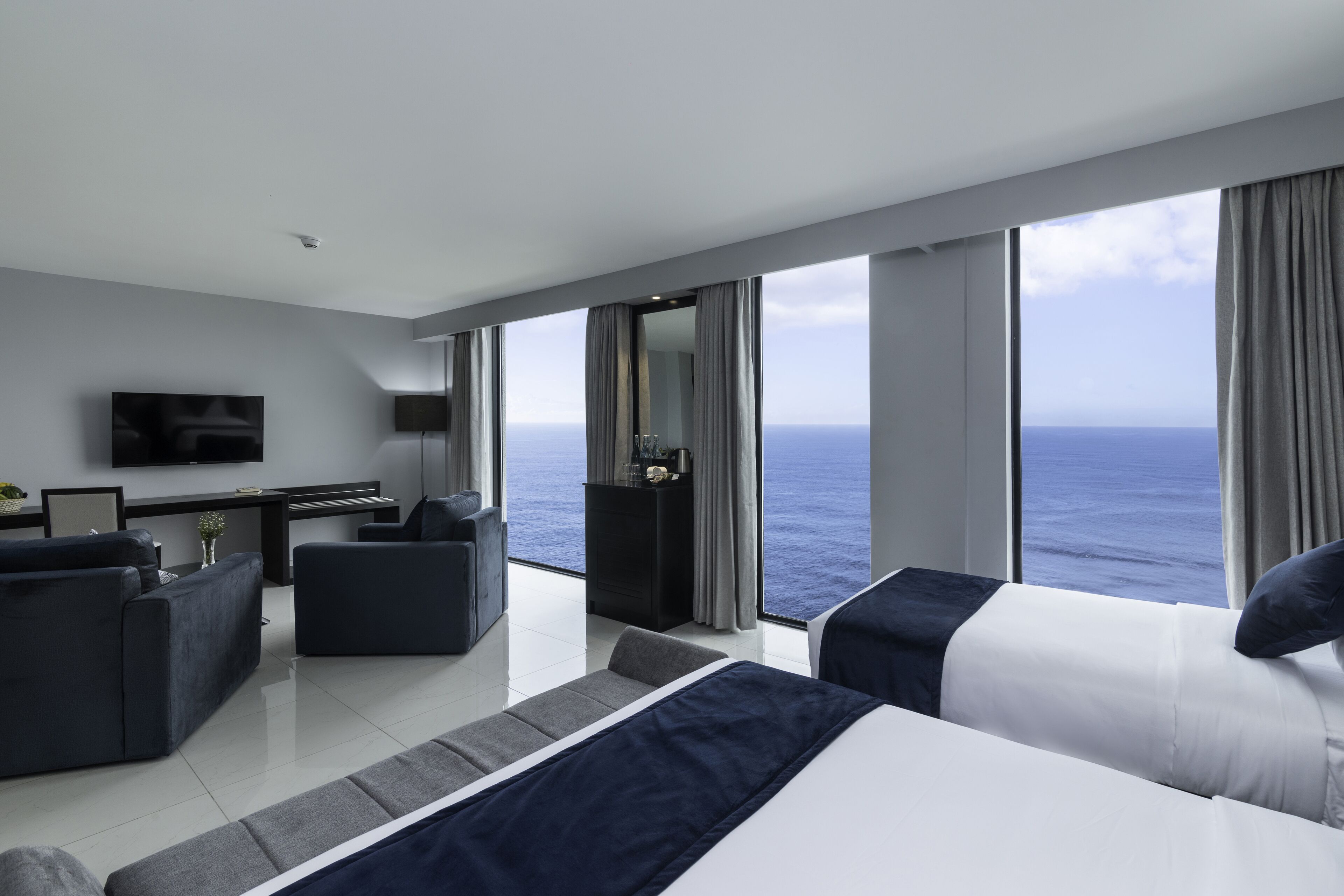 Ett modernt hotellrum med ett stort fönster som erbjuder utsikt över havet, en säng, ett skrivbord och en sittgrupp.