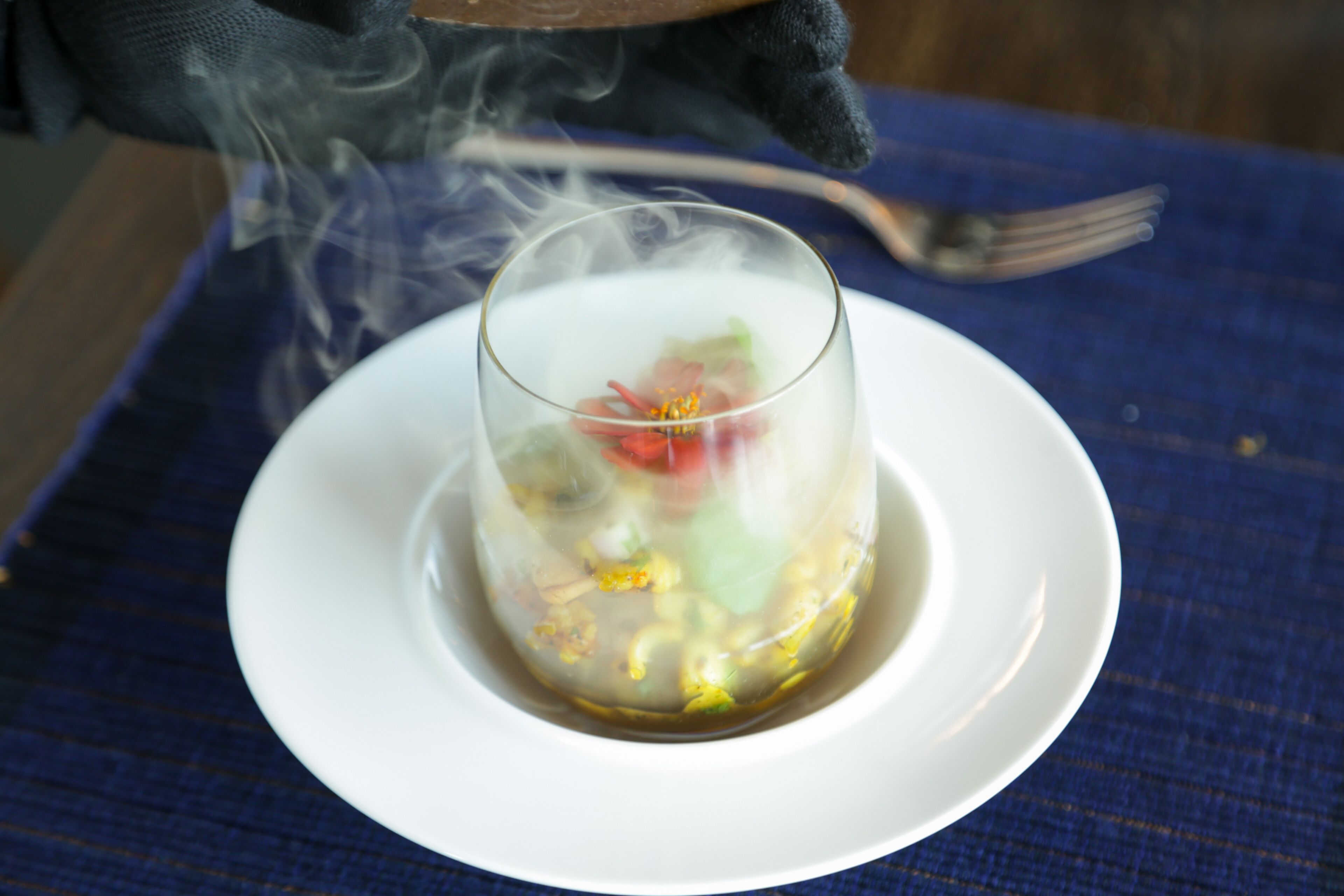 A clear glass bowl with a dessert on a white plate, placed on a blue tablecloth.