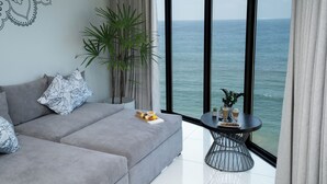 A modern living room with a grey sofa, patterned cushions, a side table with drinks, a potted plant, and a large window overlooking the sea. - MaRadha Colombo (Colombo)