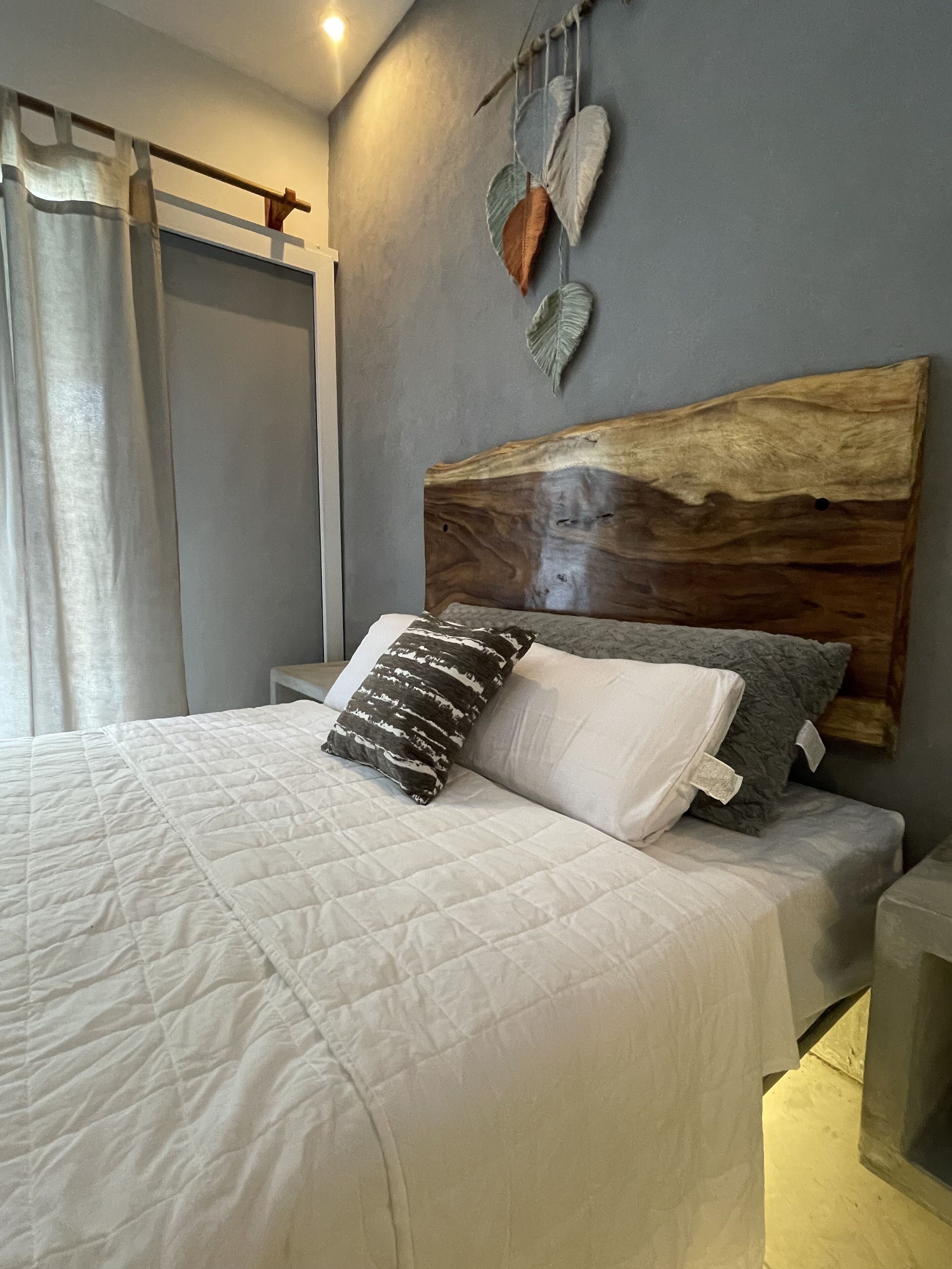 Un lit avec une couette blanche matelassée, deux oreillers, une tête de lit en bois et une décoration murale en forme de feuille.
