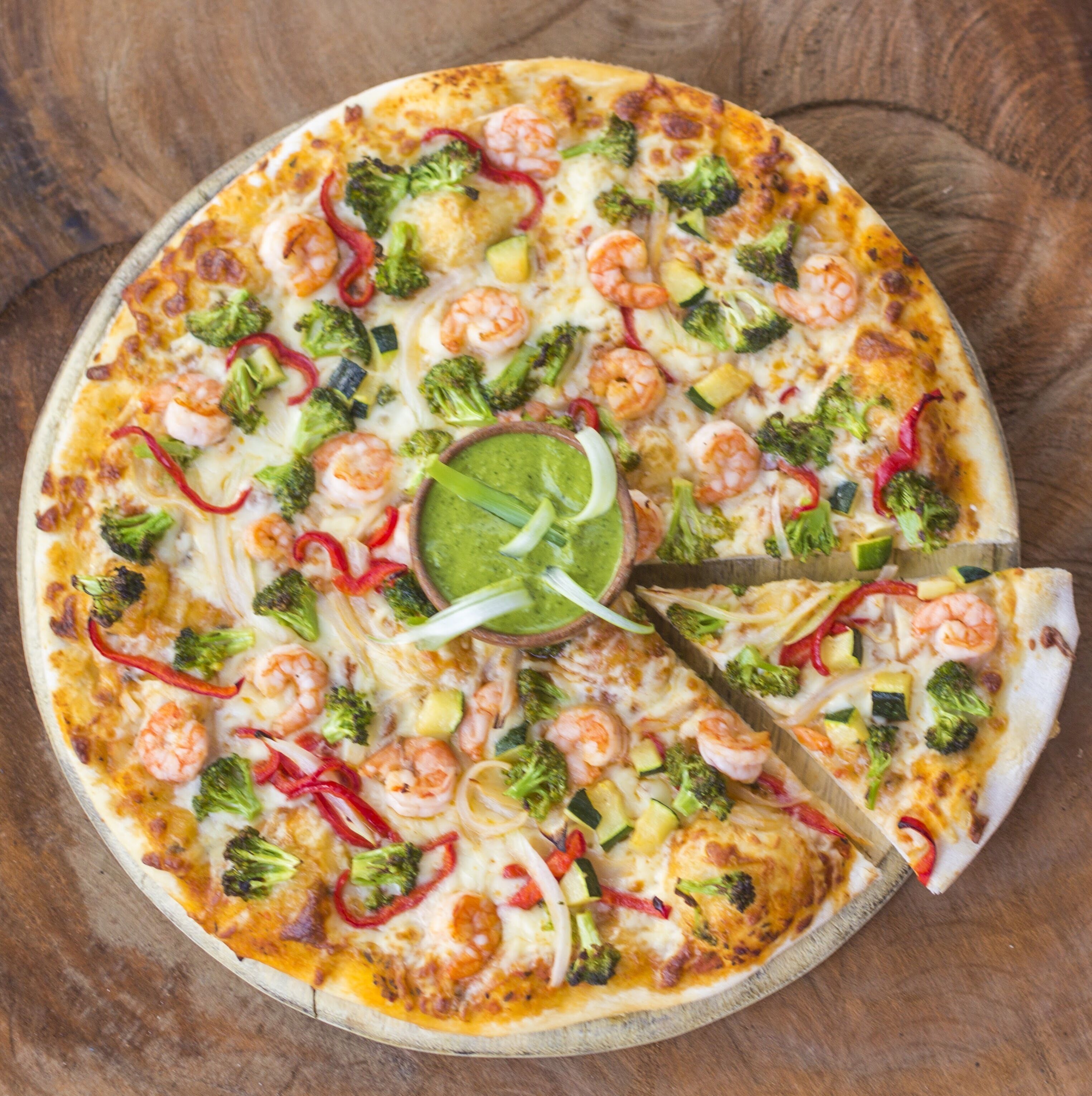 Pizza met verschillende toppings, waaronder broccoli, garnalen en kaas, op een houten plank.