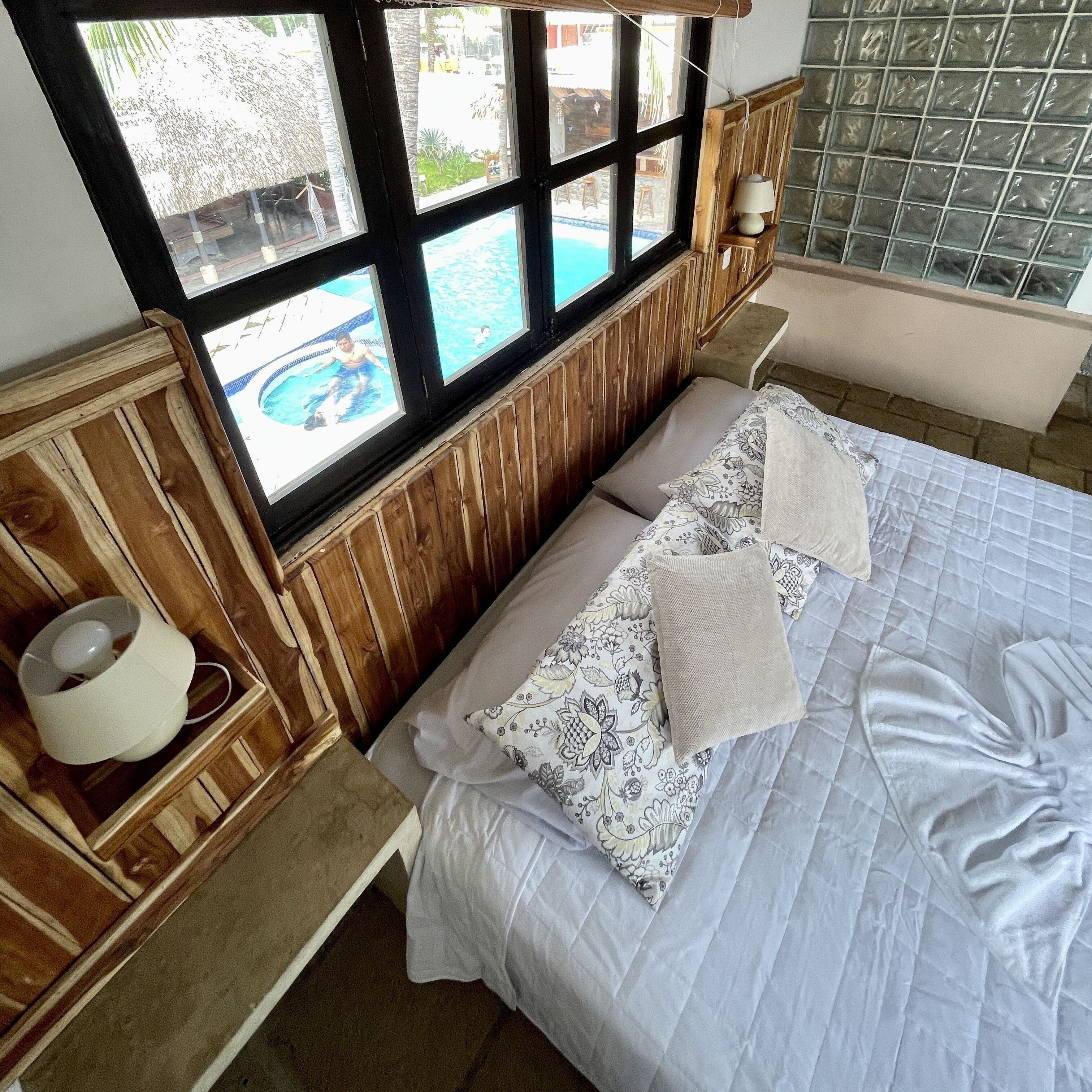 A bedroom with a large bed, a wooden headboard, a window with a view of a swimming pool, and a bedside table with a lamp.