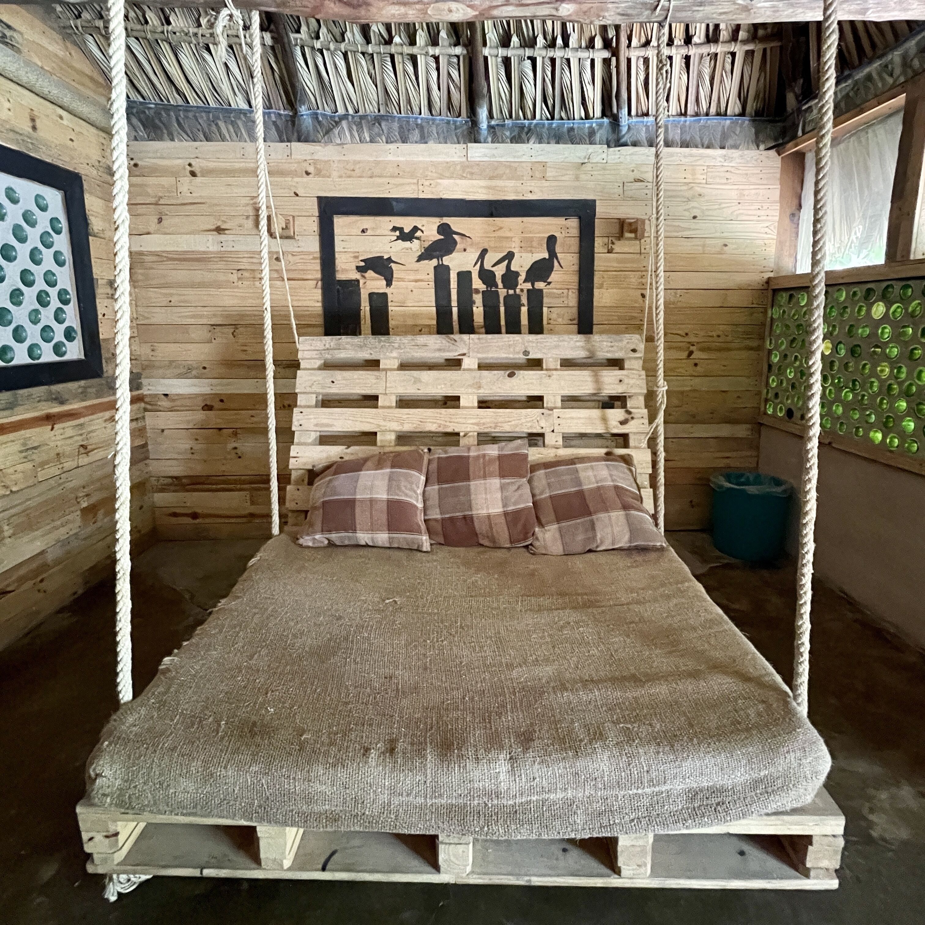 Una cama de madera rústica con dosel de cuerda, cabecera de madera con siluetas de pájaros y almohadas a cuadros.