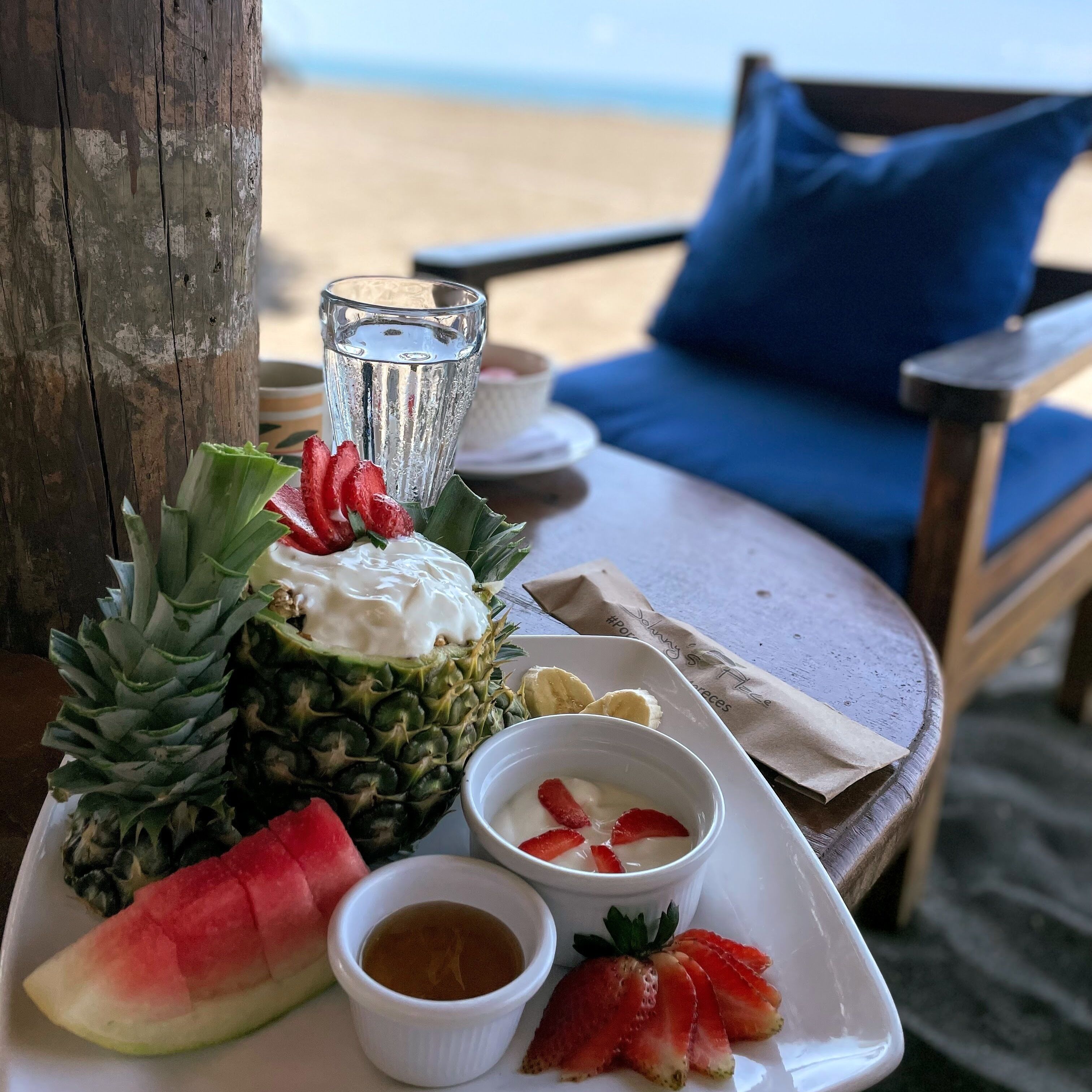 Et bord ved stranden med en tallerken mad, heriblandt ananas, vandmelon, jordbær og en lille skål sauce.