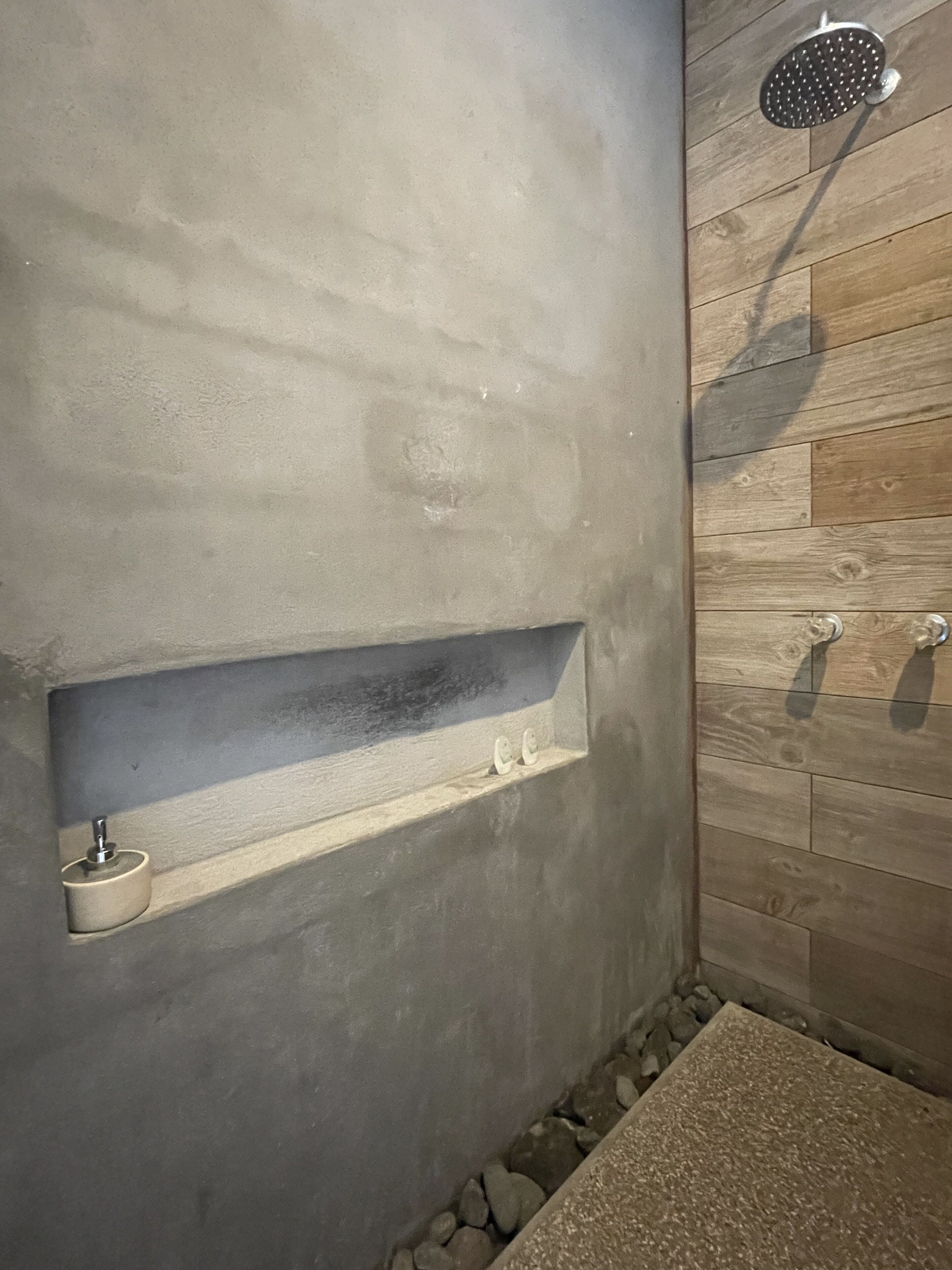 Ein Badezimmer mit einer Betonwand, einer Holzvertuschung, einer eingebauten Ablage, einem Duschkopf und einem kleinen Waschbecken.