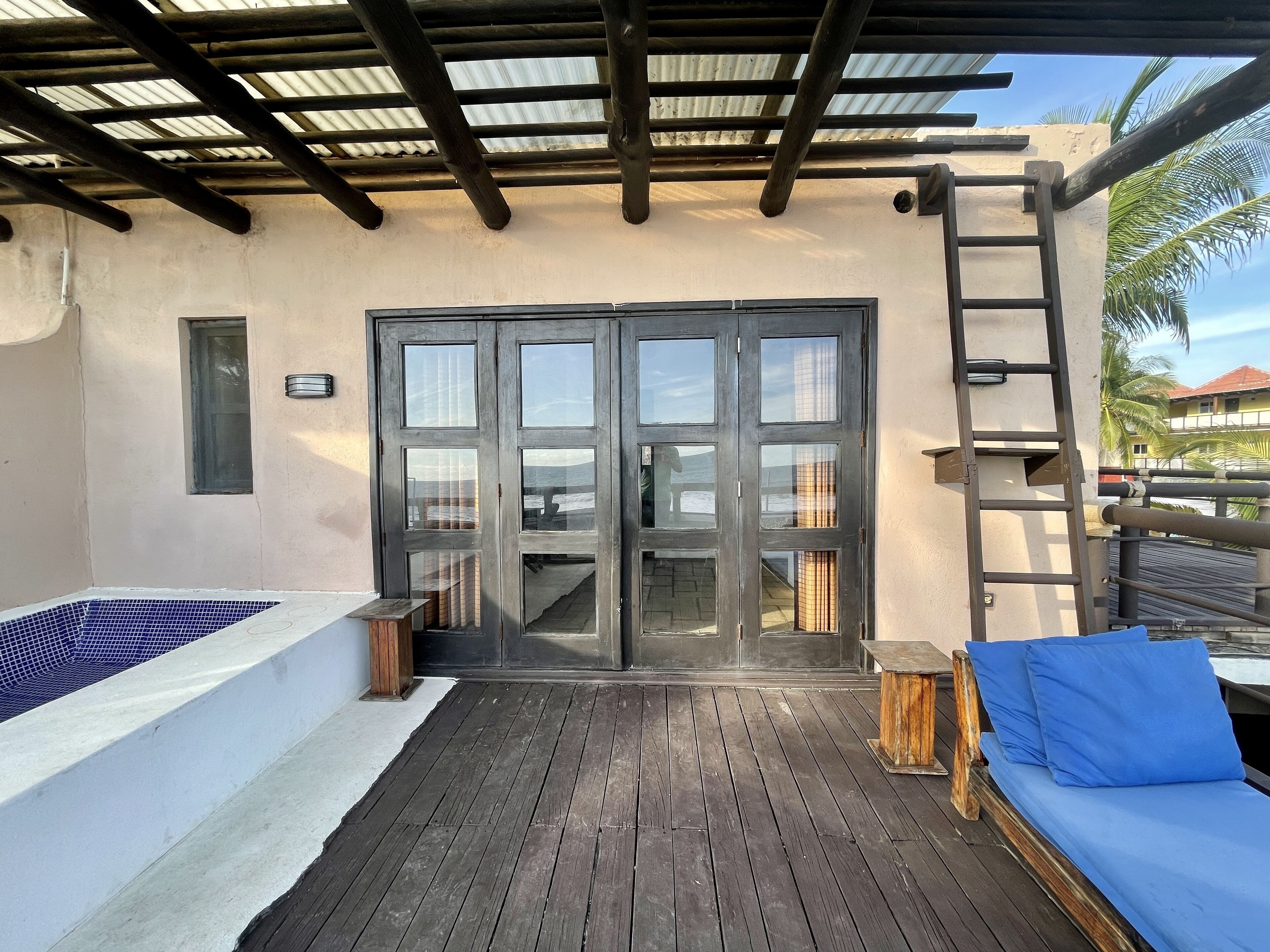 Una terraza de madera con piscina, puerta corrediza de vidrio, escalera y un banco acolchado azul.
