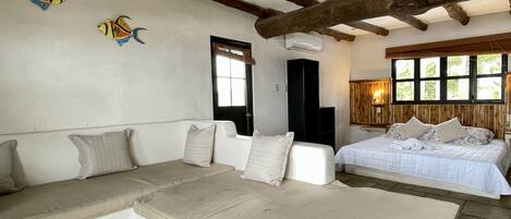 Une pièce avec un grand lit, un canapé et une petite table. Le plafond est orné de poutres en bois et une fenêtre offre une vue sur les arbres.