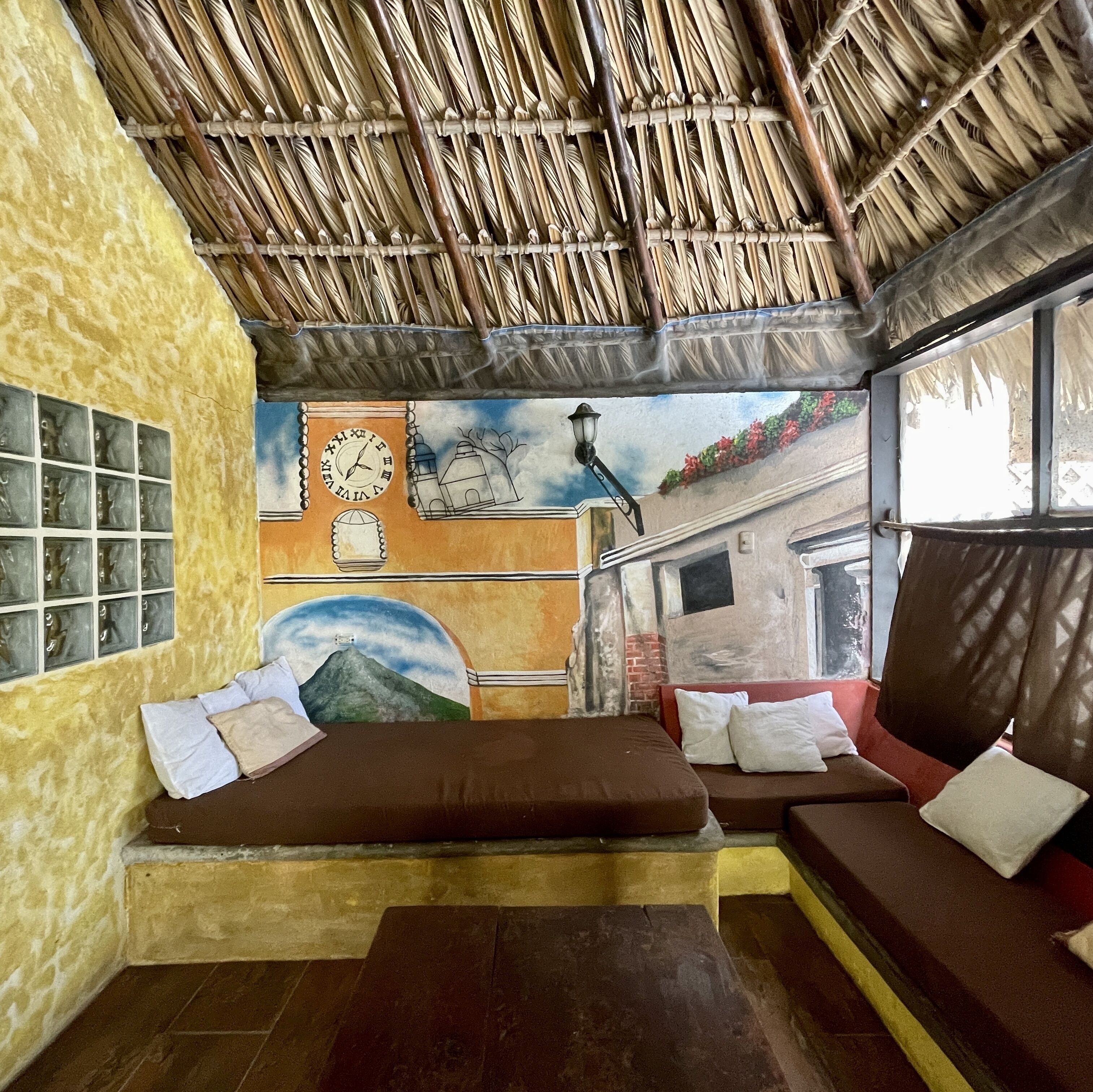 A room with a thatched roof, a mural on the wall, a bed with pillows, and a window with a view of a mountain.