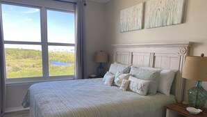 Una camera da letto con un'ampia finestra, un letto con molti cuscini, un comodino con una lampada e opere d'arte alle pareti.