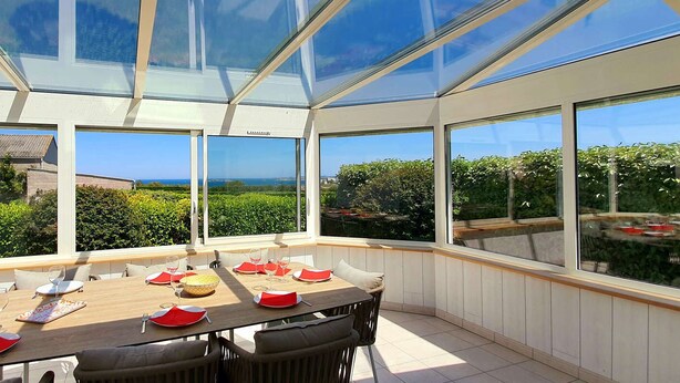 Une véranda équipée d’une table à manger pour six personnes, de grandes fenêtres et offrant une vue sur la verdure et les bâtiments.