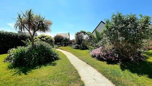 Un sentier qui traverse un jardin où l’on trouve différentes espèces de plantes, avec un bâtiment visible au loin.