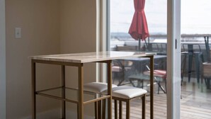 A modern room with a glass table and two chairs, a large window with a view of outdoor seating, and a red umbrella visible outside.