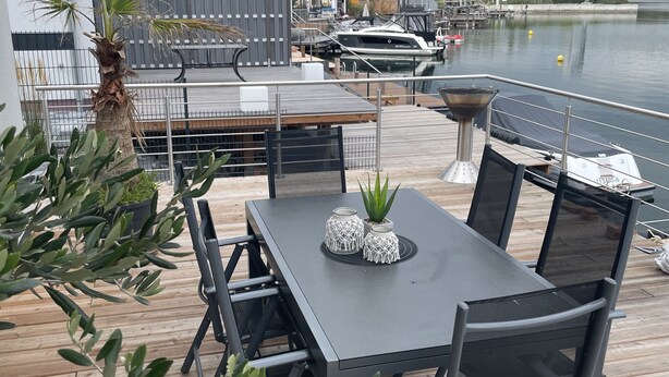 Une terrasse en bois avec une table et des chaises, donnant sur une marina où sont amarrés des bateaux et sur un lac.