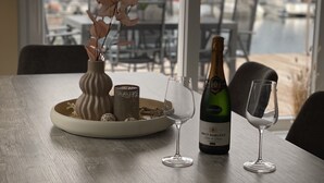 Une table à manger avec une bouteille de vin Brut Darigent, deux verres à vin, un vase contenant des fleurs séchées et une assiette de fruits secs.