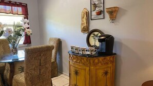 Ein Essbereich mit Tisch für vier Personen, umgeben von gemusterten Stühlen, einer Dekokabine mit Kaffeemaschine und Wandbildern.