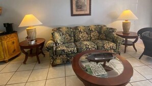 Ein Wohnzimmer mit einem blumigen Sofa, zwei Nachttischen mit Lampen, einer Kaffeemaschine, einem Holzschrank und einem Couchtisch mit Glasplatte.