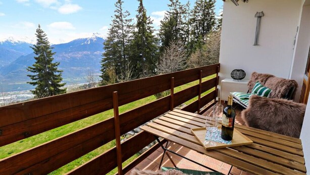 Uma varanda com um banco de madeira, uma mesa com garrafa e copos, e vista para montanhas e árvores.