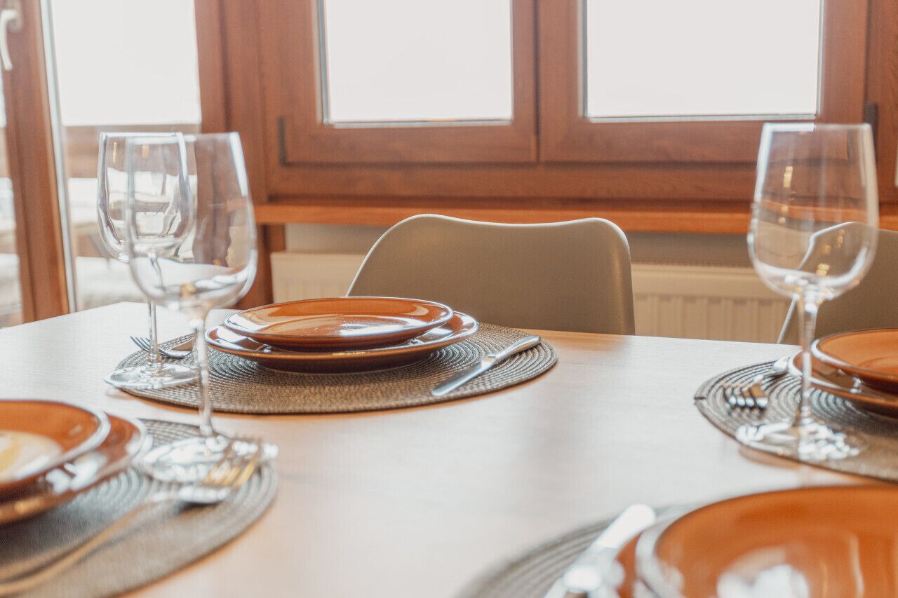 A dining table set with plates, cutlery, and glasses, with a window in the background.