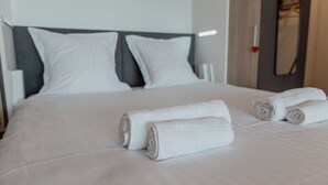 Cama bem arrumada com lençóis brancos e toalhas dobradas, cabeceira cinzenta e um espelho na parede.