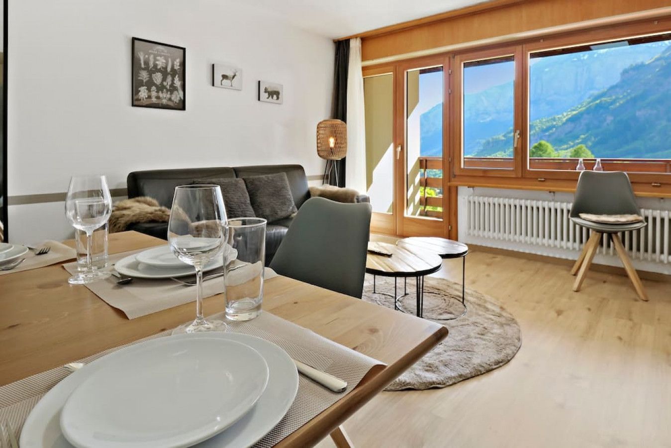 A dining area with a wooden table set for two, a sofa, and a view of mountains through large windows.