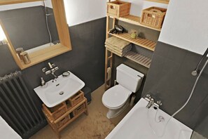 A compact bathroom with a white sink, a toilet, and a bathtub. There are wooden crates under the sink and a shelf with baskets above the toilet. - New studio with a breathtaking view of the valley (Leukerbad)