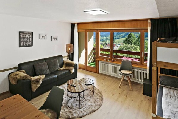 A cozy living room with a black leather sofa, a round coffee table, and a view of a scenic landscape through large windows. - New studio with a breathtaking view of the valley (Leukerbad)