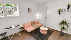 Una sala moderna con un sofá, una mesa de centro, una mesa de comedor y una pequeña mesita auxiliar. Hay una lámpara en la pared, un cuadro enmarcado y una planta en maceta.