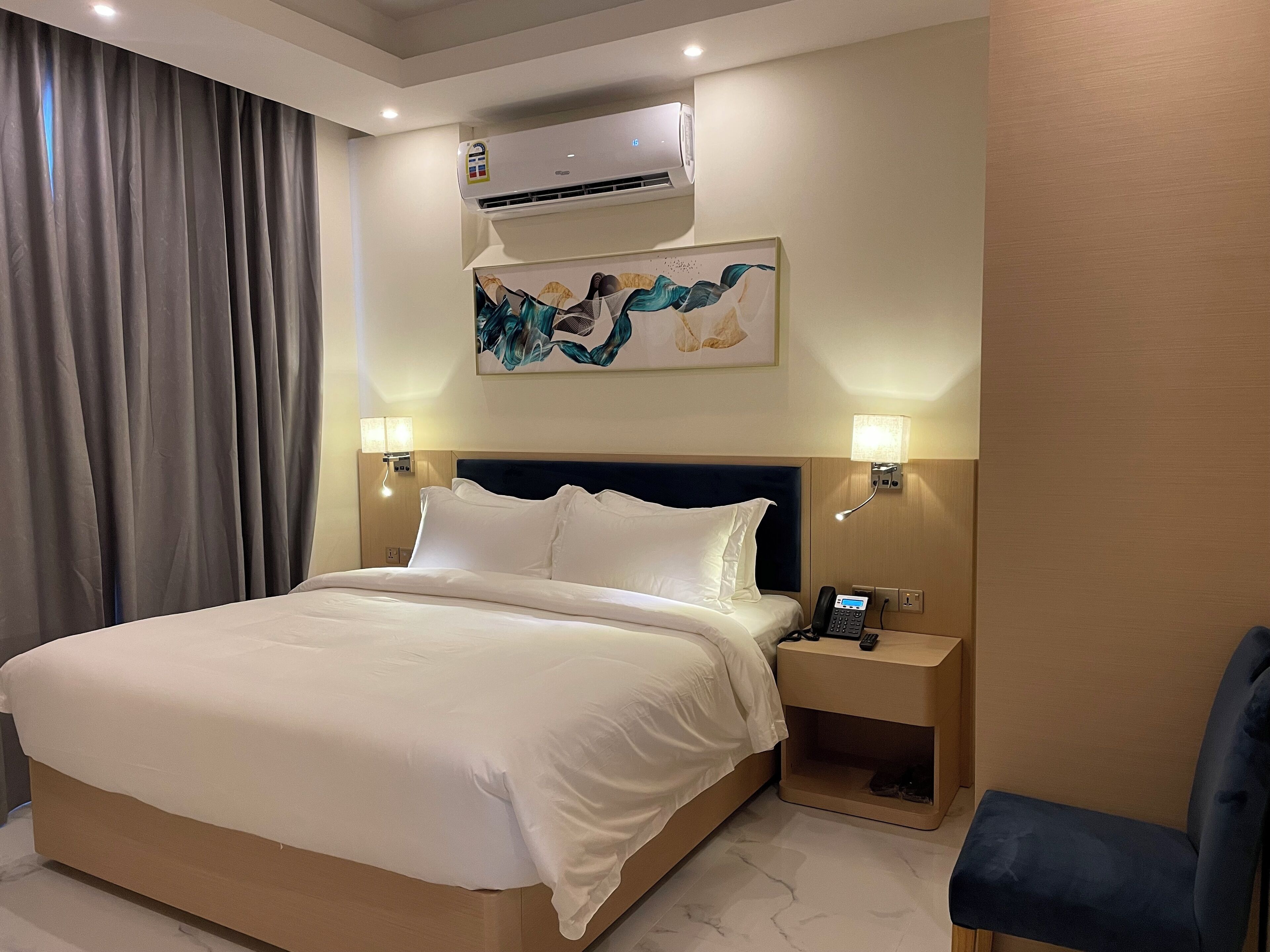 A hotel room with a large bed, a nightstand with a phone, an air conditioning unit, and a painting on the wall.