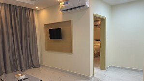 Habitación de hotel con una televisión de pantalla plana montada en la pared, una mesita con un control remoto, una cortina y una puerta que da a un dormitorio.