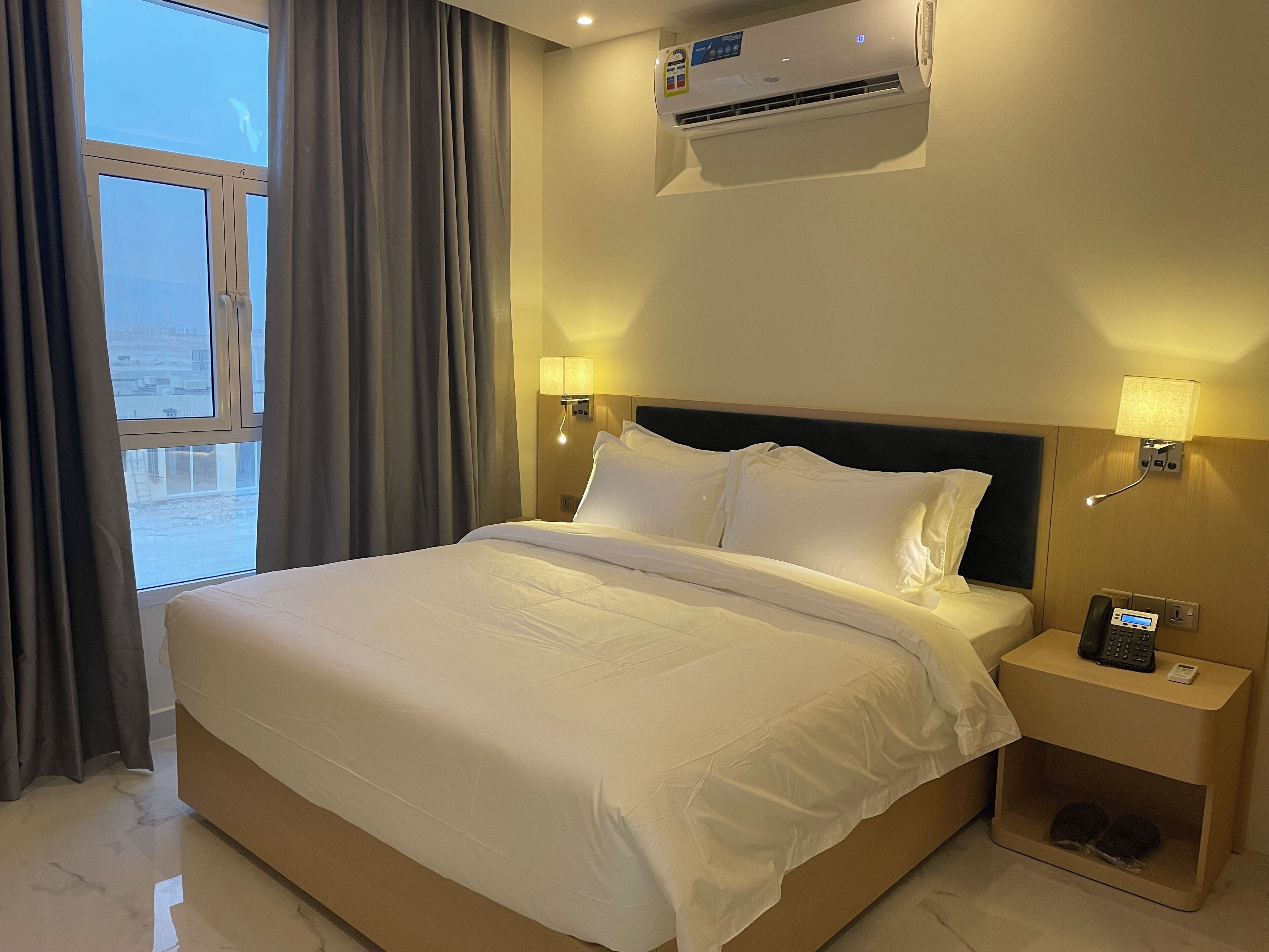 A hotel room with a large bed, two bedside lamps, a nightstand with a phone, and an air conditioning unit.