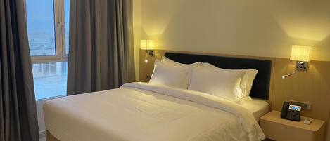 A hotel room with a large bed, two bedside lamps, a nightstand with a phone, and an air conditioning unit.