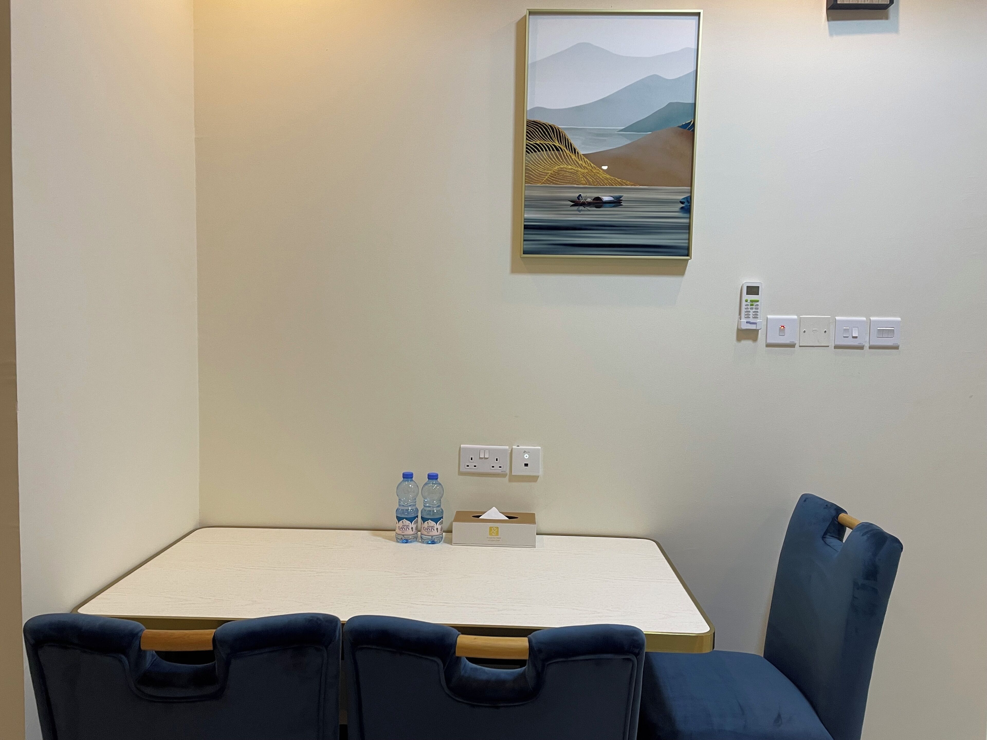 Quarto de hotel com uma mesa, duas cadeiras, um quadro e uma tomada com telefone e termostato.