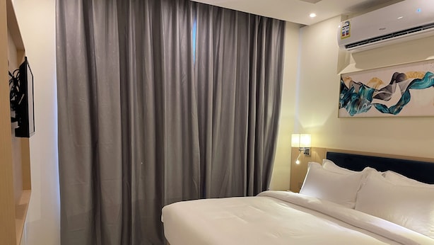 Una habitación de hotel con una cama, cortinas grises, un cuadro, un aire acondicionado y una mesita de noche con una lámpara.