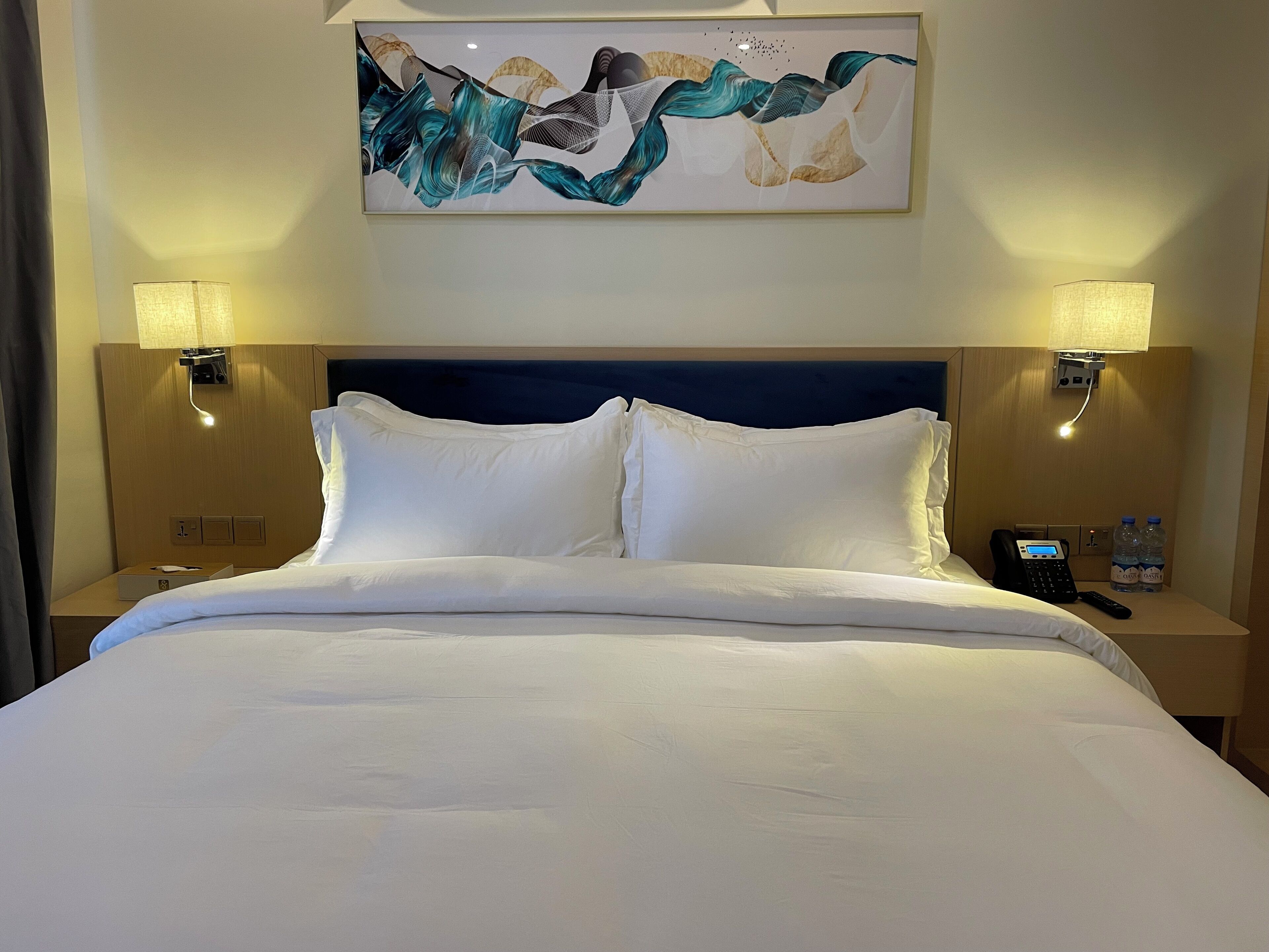 A hotel room with a large bed, two bedside lamps, a nightstand with a phone and water bottles, and a painting above the bed.