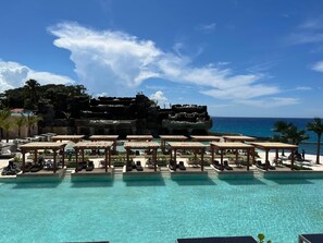 Zwembadgedeelte van een resort met meerdere ligbedden en parasols, een helder uitzicht op de oceaan en een rotsachtige klif op de achtergrond.