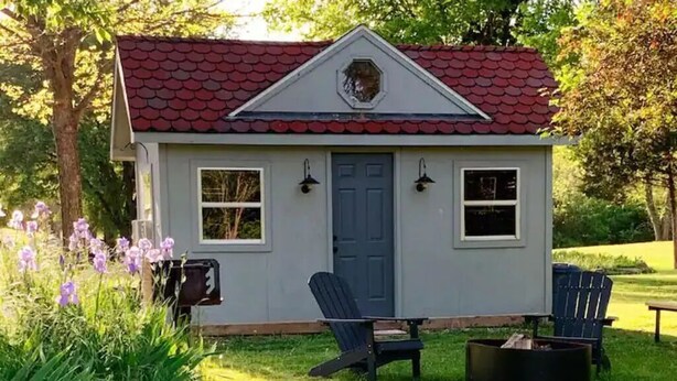 A small, single-story cabin with a red-tiled roof, a blue door, and two windows on the front. There are two Adirondack chairs and a fire pit in front of the cabin.