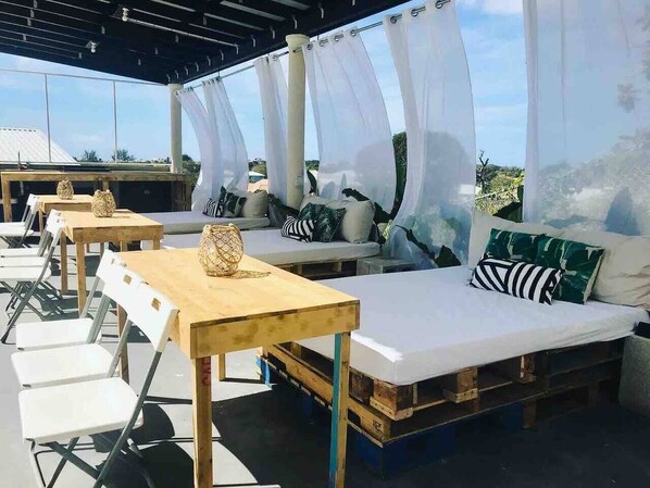 An outdoor seating area with wooden tables and white chairs, a sofa with patterned cushions, and a mural on the wall.