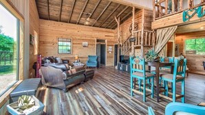 Une cabane en bois spacieuse avec une chambre à coucher en mezzanine, un coin repas avec des chaises turquoise et un salon confortable doté d’un canapé en cuir et d’une cheminée.
