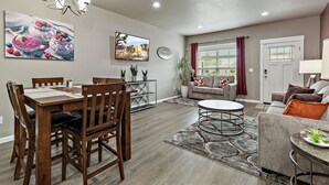A dining area with a wooden table and chairs, a sofa, and a TV mounted on the wall. - Galaxy-Themed, Fastest WiFi, 3 King Beds Home (Medford)