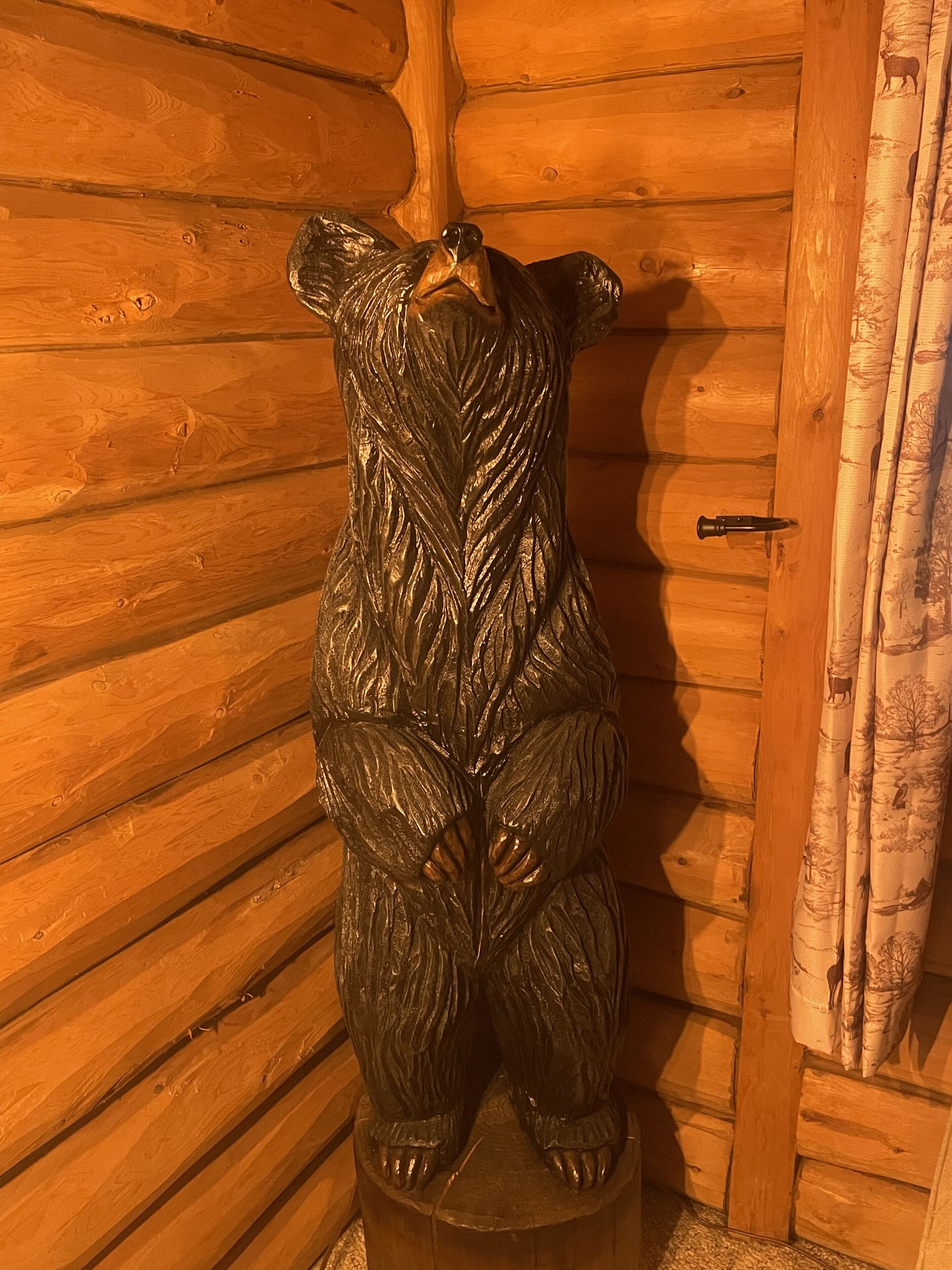 A wooden bear sculpture mounted on a wooden post in a cabin with a wooden door and curtains.