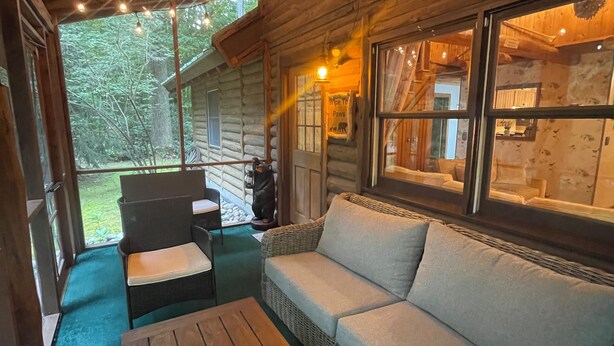 Interior acogedor de una cabaña de troncos con techo de madera, luces colgantes, un sofá de mimbre, una mesa de madera y vistas del bosque circundante.