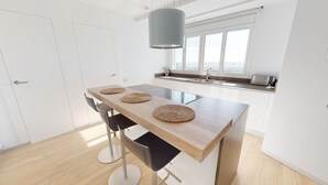 A modern kitchen with a wooden table, two chairs, a hanging light fixture, a window with a view, and a built-in oven. - APTO PANORAMIC (Ampolla)